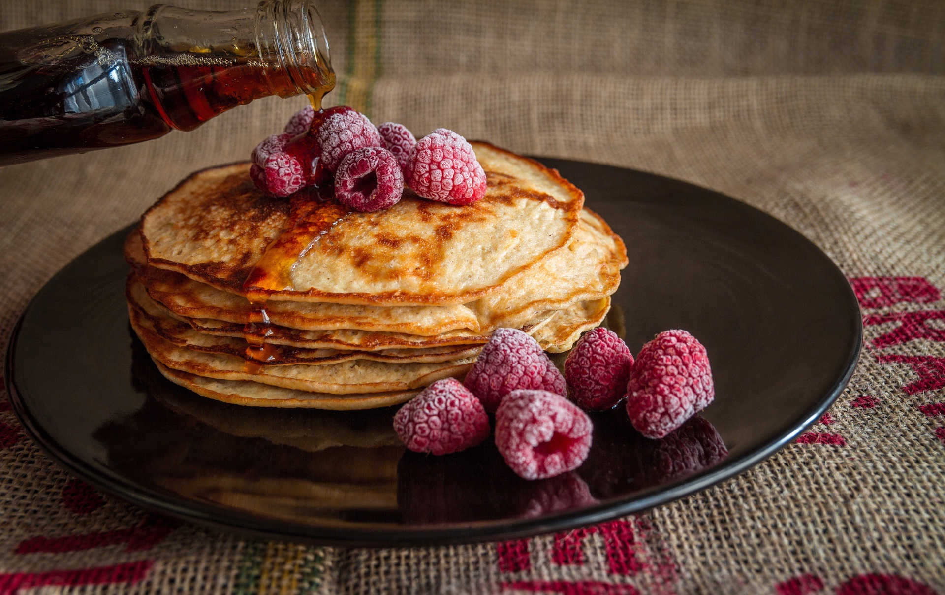 Maple syrup being poured on stack of pancakes with raspberries